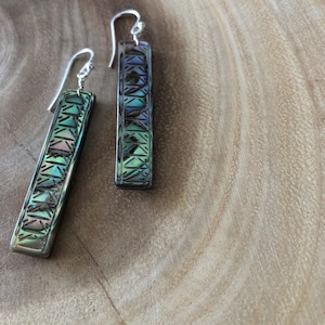 May include: A pair of silver dangle earrings with a geometric pattern made of iridescent abalone shell. The earrings are hanging on a wooden surface.
