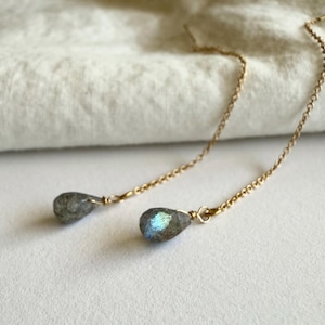 Puede incluir: Un par de pendientes de cadena dorada con piedras preciosas en forma de lágrima, grises y azules iridiscentes. Los pendientes se muestran sobre una superficie blanca con una tela suave de color blanquecino en el fondo.