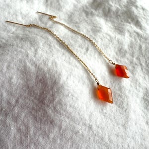 Puede incluir: Un par de pendientes de cadena dorados. Cada pendiente presenta una pequeña gema facetada de color rojo anaranjado en forma de diamante. Los pendientes se muestran sobre una superficie blanca texturizada.