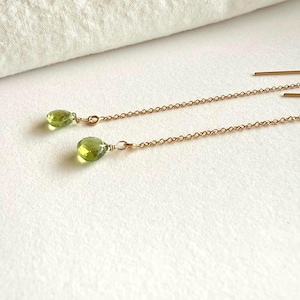 May include: A pair of delicate gold-colored chain earrings, each featuring a pear-shaped, light green gemstone. The earrings are displayed on a white surface, with a soft, white fabric in the background.