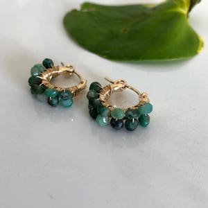 May include: Gold hoop earrings with green gemstone beads. The earrings are made of gold wire and have a cluster of green beads hanging from each hoop.