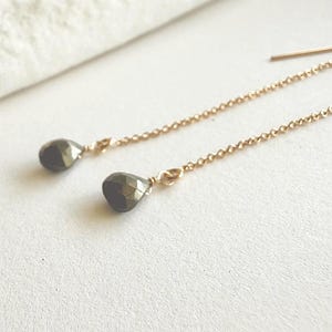 May include: A pair of gold-colored chain earrings with teardrop-shaped, dark gray pyrite stones. The earrings are displayed on a white surface, with a soft, off-white fabric in the background.