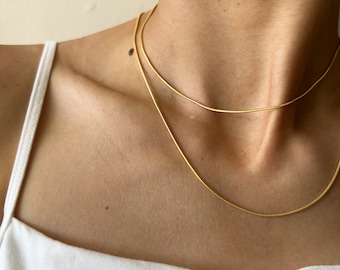 Könnte beinhalten: Zwei goldene Kettenhalsbänder, die übereinander auf dem Hals einer Person liegen. Die Halsbänder sind dünn und zart und beide aus glänzendem Goldmetall.