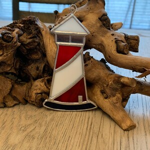 May include: A stained glass lighthouse ornament with red, white, and blue accents. The lighthouse is sitting on a piece of driftwood.