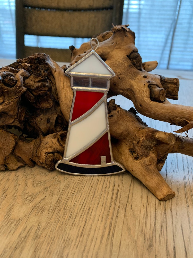May include: A stained glass lighthouse ornament with red, white, and blue accents. The lighthouse is sitting on a piece of driftwood.