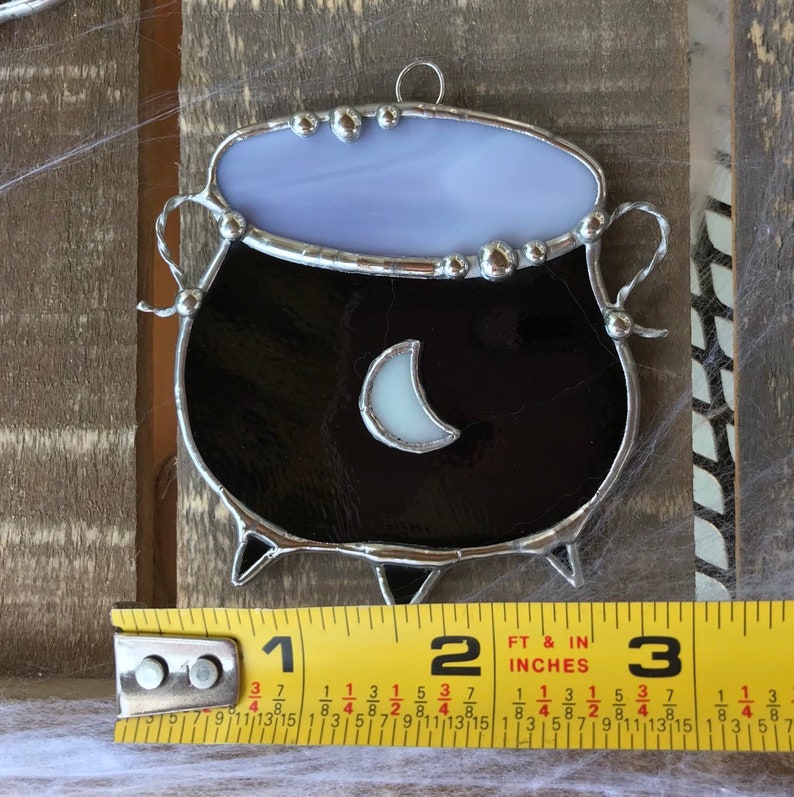 May include: A stained glass cauldron ornament with a crescent moon design. The cauldron is black with a silver rim and a light purple top. The ornament is approximately 2 inches tall.