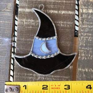 May include: A stained glass witch hat ornament with a crescent moon design. The hat is black with a purple brim and silver accents. The moon is white with a silver crescent.