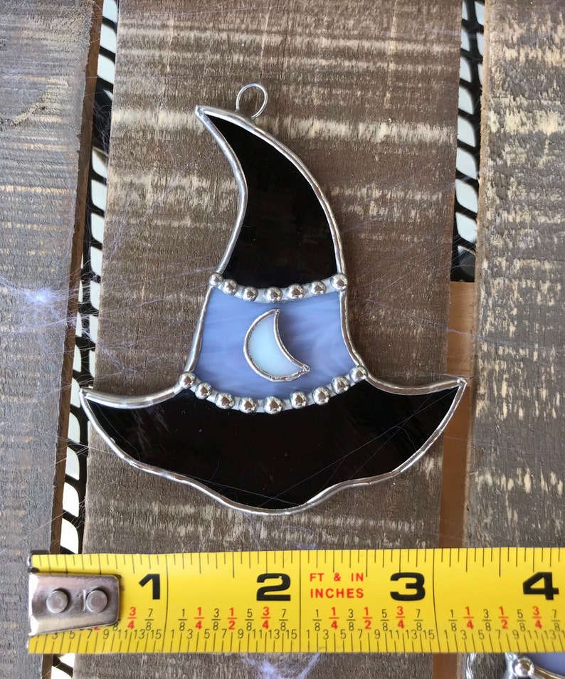 May include: A stained glass witch hat ornament with a crescent moon design. The hat is black with a purple brim and silver accents. The moon is white with a silver crescent.