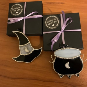 May include: Two stained glass Halloween ornaments, a black witch hat with a white crescent moon and a black cauldron with a white crescent moon, are displayed on a wooden surface. Each ornament is packaged in a black gift box with a purple ribbon and a "Handmade with Love" sticker.