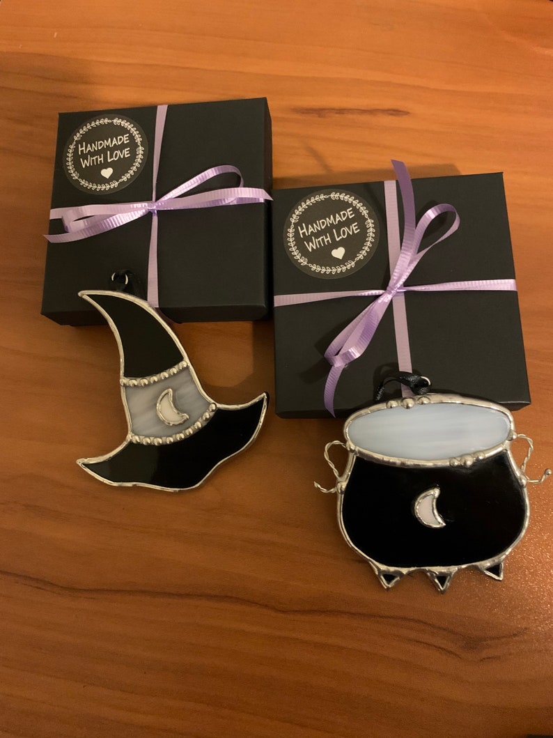 May include: Two stained glass Halloween ornaments, a black witch hat with a white crescent moon and a black cauldron with a white crescent moon, are displayed on a wooden surface. Each ornament is packaged in a black gift box with a purple ribbon and a "Handmade with Love" sticker.