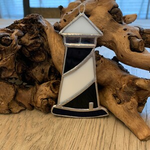 May include: A stained glass lighthouse ornament with black and white stripes. The lighthouse is made of glass and has a silver frame. It is hanging from a silver loop.
