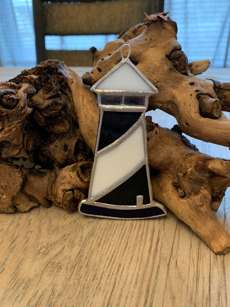 May include: A stained glass lighthouse ornament with black and white stripes. The lighthouse is made of glass and has a silver frame. It is hanging from a silver loop.