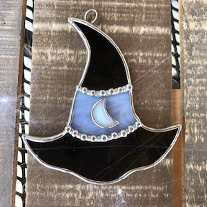May include: A stained glass witch hat ornament with a black brim, a purple and white body, and a silver crescent moon. The hat is decorated with silver beads around the brim.