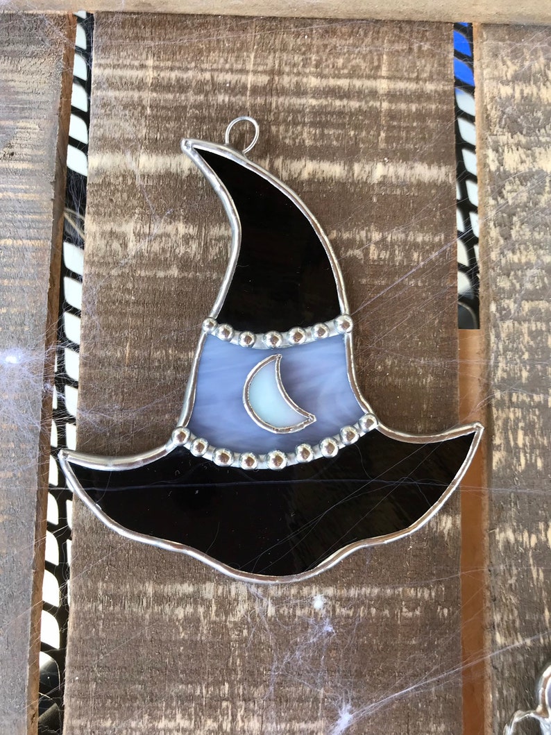 May include: A stained glass witch hat ornament with a black brim, a purple and white body, and a silver crescent moon. The hat is decorated with silver beads around the brim.