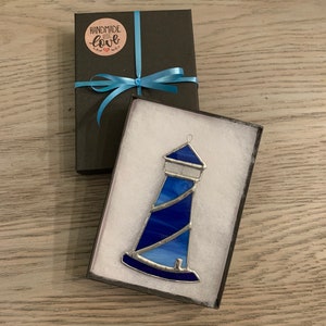 May include: A blue stained glass lighthouse ornament in a white gift box with a black lid. The lid has a blue ribbon and a round sticker that says "Handmade with love".