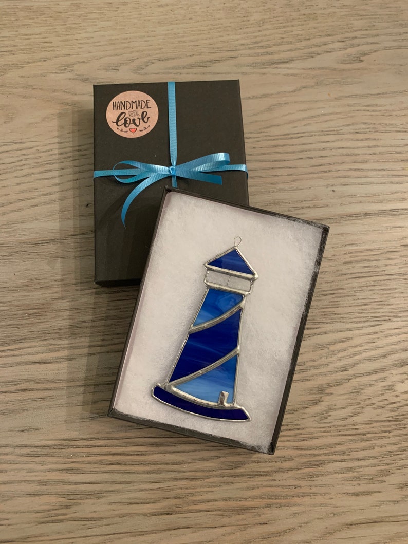May include: A blue stained glass lighthouse ornament in a white gift box with a black lid. The lid has a blue ribbon and a round sticker that says "Handmade with love".