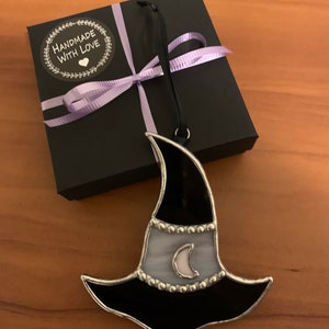 May include: A black and white stained glass witch hat ornament with a crescent moon design. The ornament is packaged in a black gift box with a purple ribbon and a "Handmade with Love" sticker.