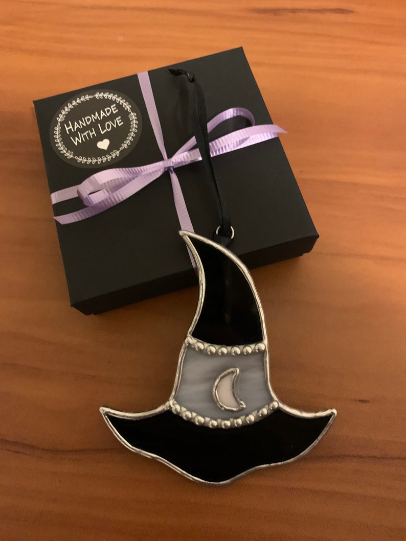 May include: A black and white stained glass witch hat ornament with a crescent moon design. The ornament is packaged in a black gift box with a purple ribbon and a "Handmade with Love" sticker.