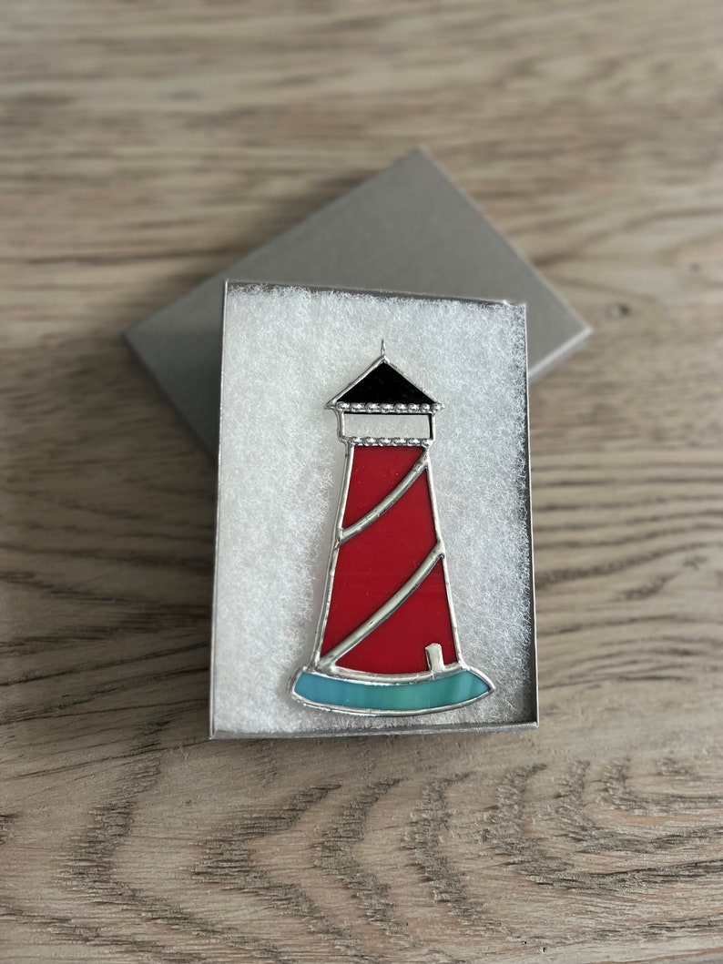 May include: A red and white lighthouse with a black roof and a blue base, in a white gift box.