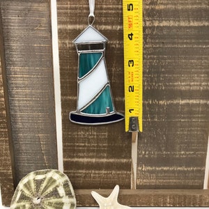 May include: A stained glass lighthouse ornament with a white and teal design. The ornament is hanging from a white ribbon and is measured against a yellow measuring tape.