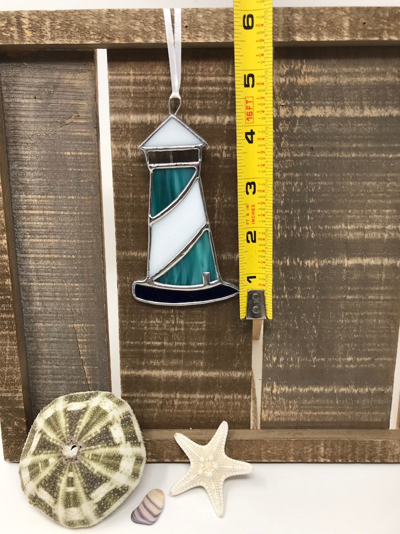 May include: A stained glass lighthouse ornament with a white and teal design. The ornament is hanging from a white ribbon and is measured against a yellow measuring tape.