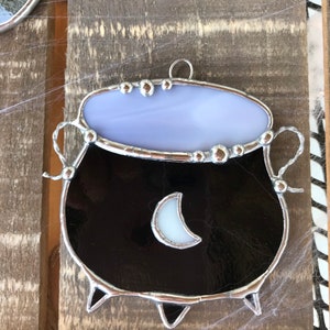 May include: A stained glass cauldron ornament with a black body, a white crescent moon, and a lavender top. The cauldron has a silver metal frame and is hanging from a silver loop.