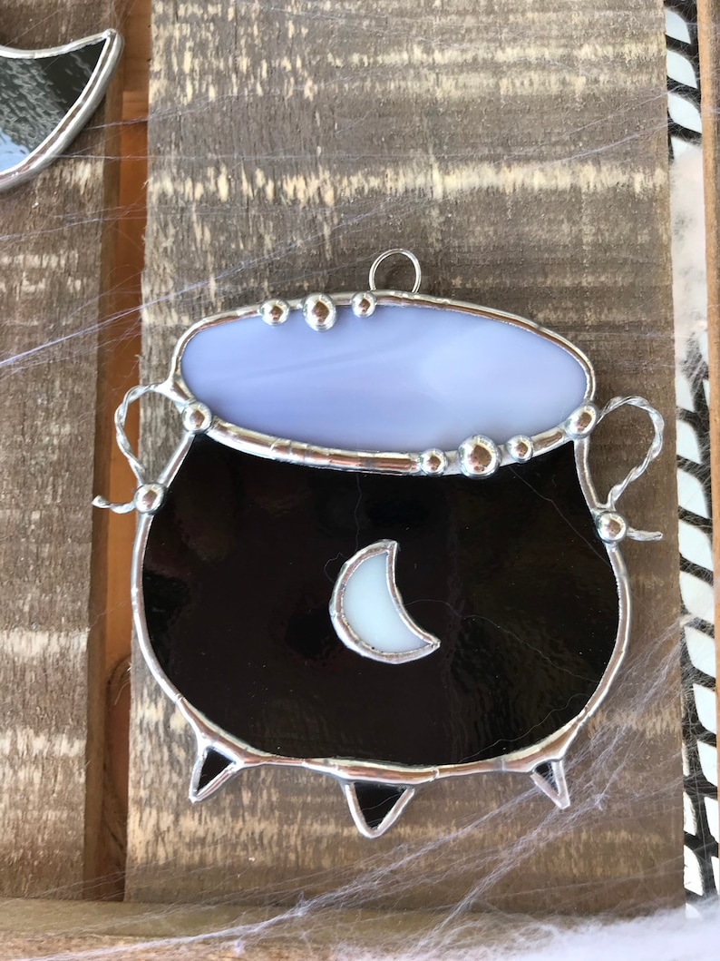 May include: A stained glass cauldron ornament with a black body, a white crescent moon, and a lavender top. The cauldron has a silver metal frame and is hanging from a silver loop.