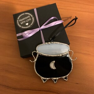 May include: A black stained glass cauldron ornament with a white crescent moon and a purple ribbon on a black gift box with a "Handmade with Love" sticker.