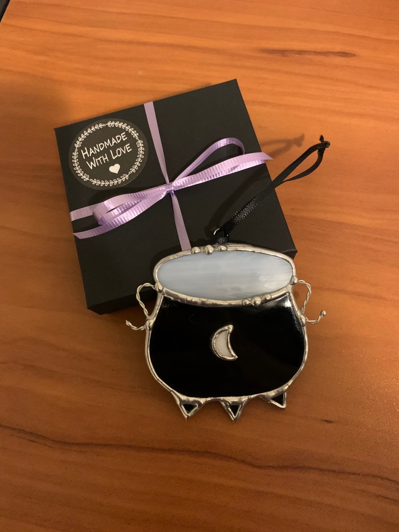 May include: A black stained glass cauldron ornament with a white crescent moon and a purple ribbon on a black gift box with a "Handmade with Love" sticker.