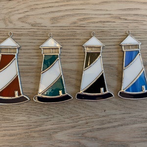 May include: Four stained glass lighthouse ornaments in different colors. The lighthouses are white with a colored stripe and a black base. The colors are red, green, black, and blue.
