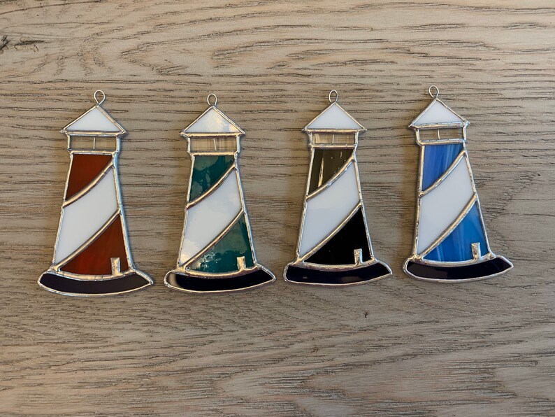 May include: Four stained glass lighthouse ornaments in different colors. The lighthouses are white with a colored stripe and a black base. The colors are red, green, black, and blue.