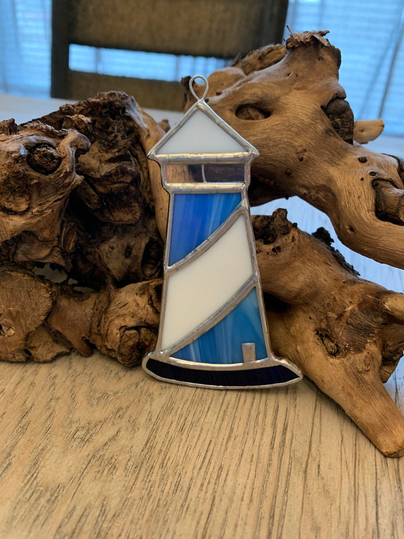 May include: A stained glass lighthouse ornament with a blue and white striped design. The lighthouse has a black base and a silver metal frame.
