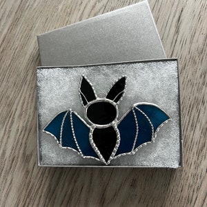 May include: A stained glass bat ornament with black and blue glass. The bat is in a white box with a silver lid.