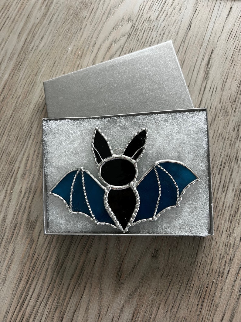 May include: A stained glass bat ornament with black and blue glass. The bat is in a white box with a silver lid.