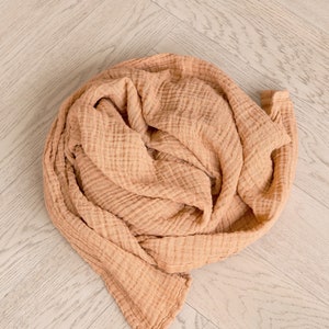 May include: A soft, peach-colored muslin swaddle blanket is folded and rolled up on a light wood floor.