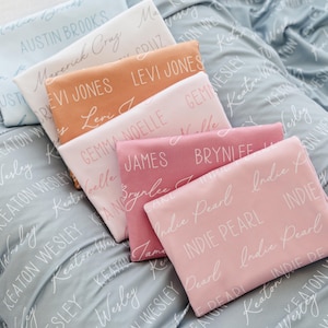 May include: A collection of personalized baby blankets in various colors, including light blue, white, peach, pink, and rose. Each blanket is folded and features a name printed in a cursive font. The background blanket is light blue with a repeating name pattern.