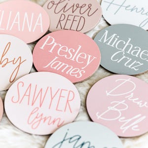 Op de afbeelding: Een verzameling ronde houten labels in roze, wit en grijs met namen geschreven in wit en roze schrift. De namen zijn Eliana, Oliver Reed, Henry, Presley James, Michael Cruz, Ruby, Sawyer Lynn, Drew Belle en Jamie.
