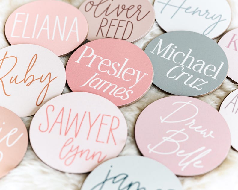 Op de afbeelding: Een verzameling ronde houten labels in roze, wit en grijs met namen geschreven in wit en roze schrift. De namen zijn Eliana, Oliver Reed, Henry, Presley James, Michael Cruz, Ruby, Sawyer Lynn, Drew Belle en Jamie.