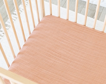 May include: A peach-colored fitted crib sheet made of a soft, textured fabric. The sheet is in a wooden crib.