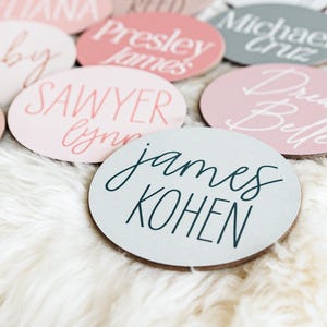 Op de afbeelding: Een verzameling ronde, houten naamschildjes met verschillende namen geschreven in witte en zwarte kalligrafie. De namen zijn "Ruby", "Sawyer Lynn", "James Kohen", "Drew Belle", "Michael Cruz" en "Presley James".