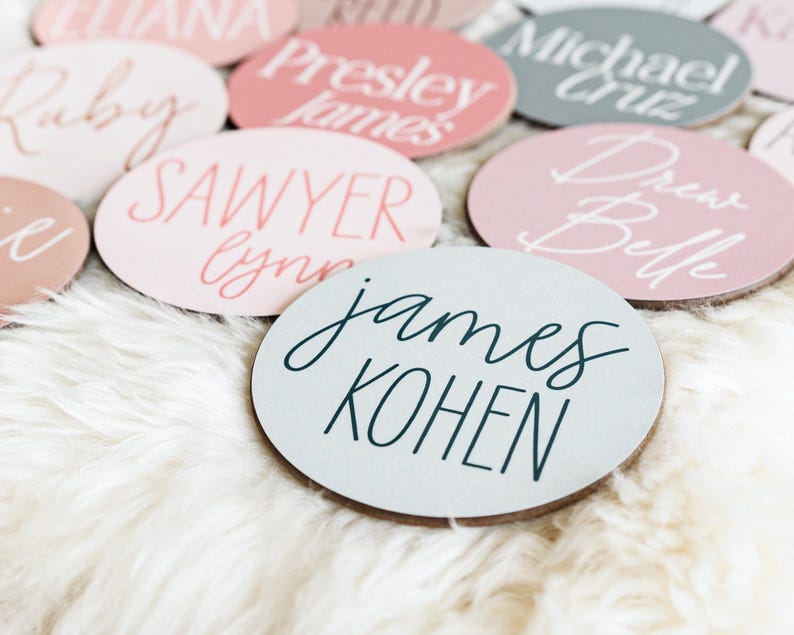 Op de afbeelding: Een verzameling ronde, houten naamschildjes met verschillende namen geschreven in witte en zwarte kalligrafie. De namen zijn "Ruby", "Sawyer Lynn", "James Kohen", "Drew Belle", "Michael Cruz" en "Presley James".