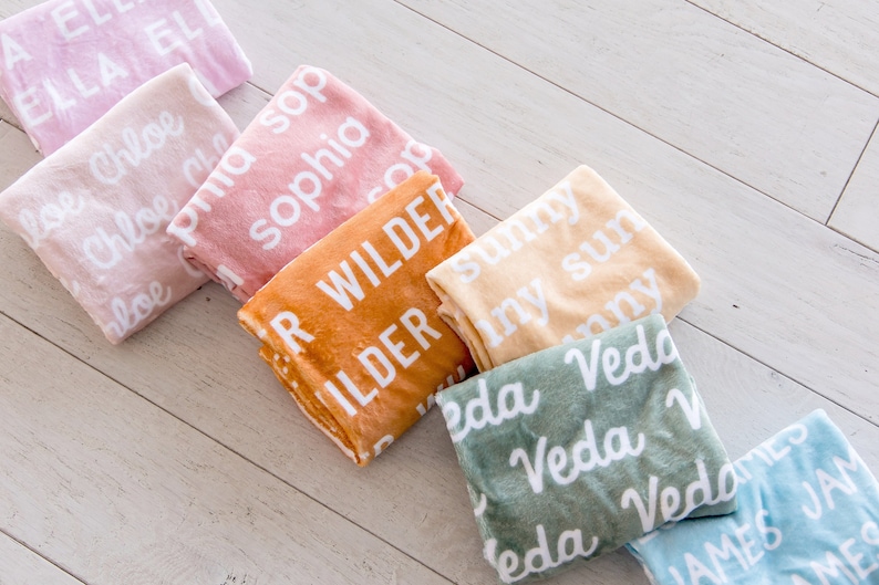 May include: A collection of personalized blankets in various pastel colors, including pink, peach, orange, yellow, green, and blue. Each blanket features a name printed in white, with names like "Ella", "Chloe", "Sophia", "Wilder", "Sunny", "Veda", and "James".