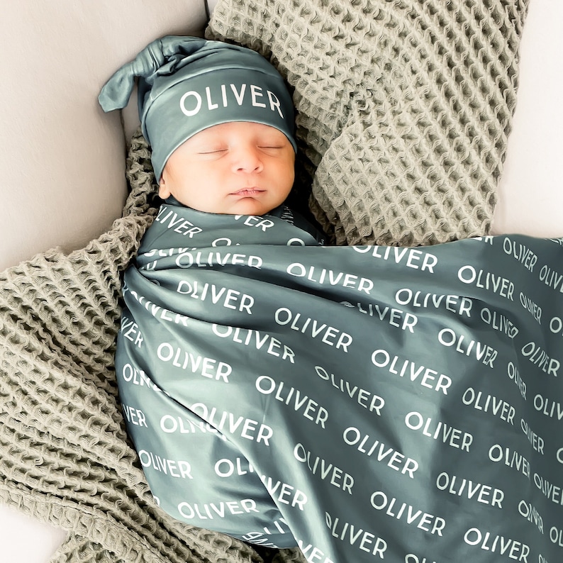 May include: A sleeping baby wrapped in a teal blanket and wearing a matching hat, both printed with the name "OLIVER" in white. The baby rests on a gray waffle-knit blanket.