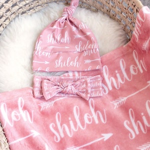 May include: A baby gift set in a woven basket, featuring a pink blanket, hat, and headband. The set is adorned with the name "Shiloh" in white script, along with arrow designs. The items are made of soft, plush material.