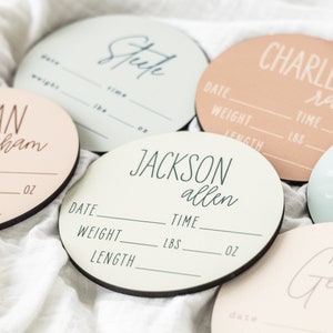 May include: Five round wooden baby announcement plaques with black borders. Each plaque has a different name and the text "Date", "Weight", "Length", "Time", "LBS", and "OZ". The names are "Fran Graham", "Steep", "Jackson Allen", "Charlee Rae", and "Gem".