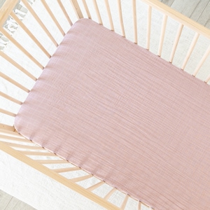 May include: A light pink fitted crib sheet made of a soft, textured fabric. The sheet is placed inside a wooden crib with a slatted design. The sheet covers the entire crib mattress.