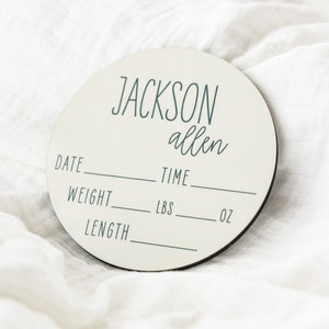 May include: A round, off-white wooden plaque with the name "JACKSON allen" written in black script. The plaque has spaces to fill in the date, time, weight in pounds and ounces, and length.