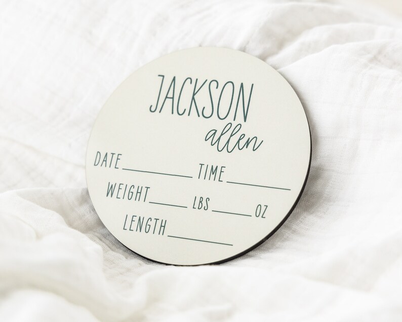 May include: A round, off-white wooden plaque with the name "JACKSON allen" written in black script. The plaque has spaces to fill in the date, time, weight in pounds and ounces, and length.