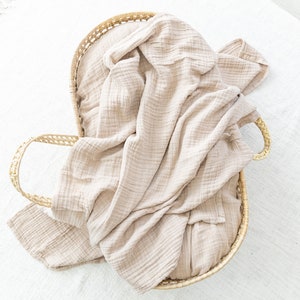 May include: A woven wicker basket with a light brown muslin blanket inside. The blanket is folded and draped over the sides of the basket.