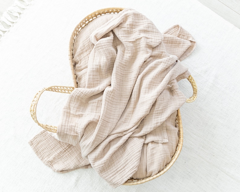 May include: A woven wicker basket with a light brown muslin blanket inside. The blanket is folded and draped over the sides of the basket.